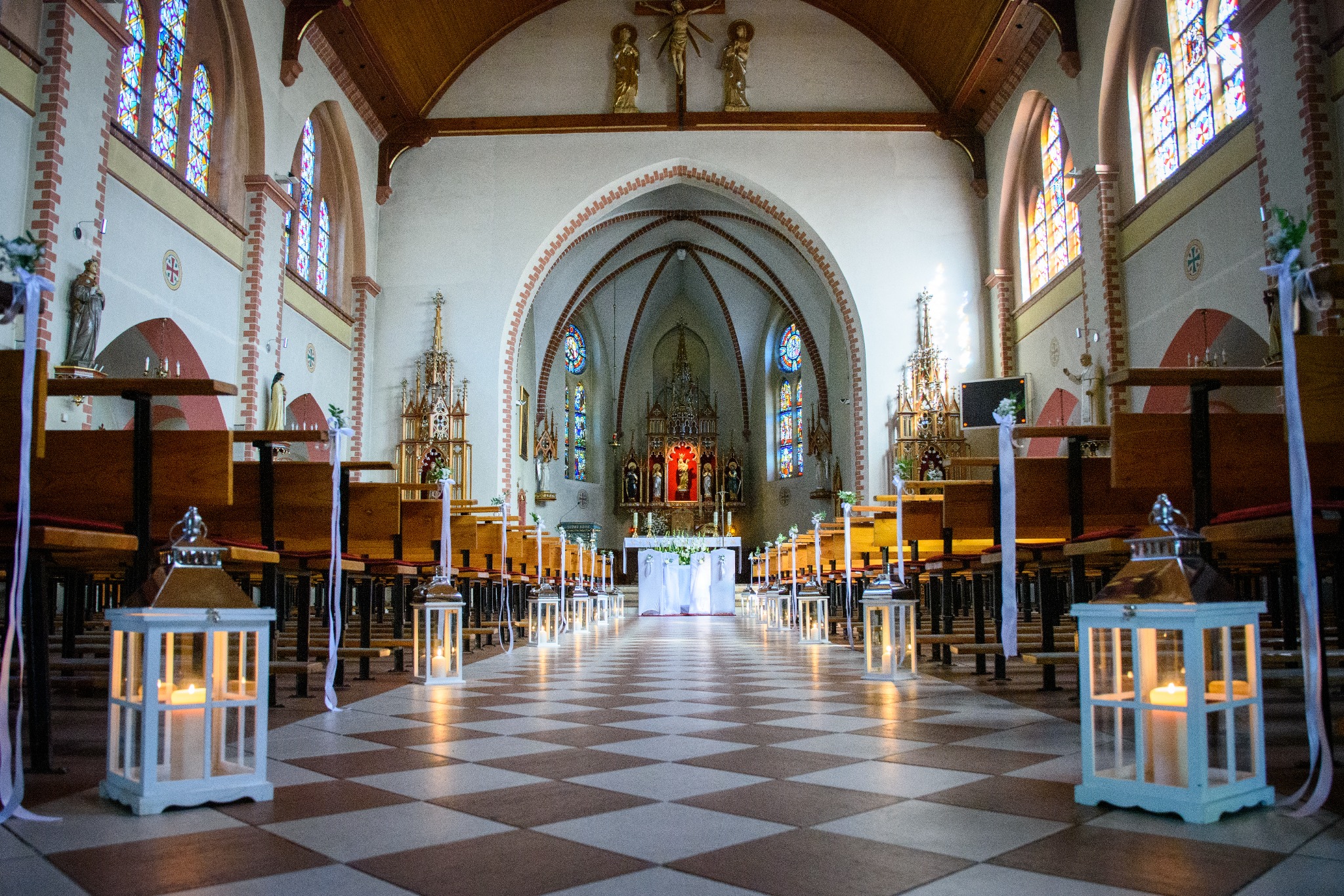 Udekorowany kościół przed ceremonią ślubną, z białymi lampionami ze świecami wzdłuż nawy głównej i białymi wstążkami na ławkach, wzorzysta podłoga, oświetlenie górne i witraże.