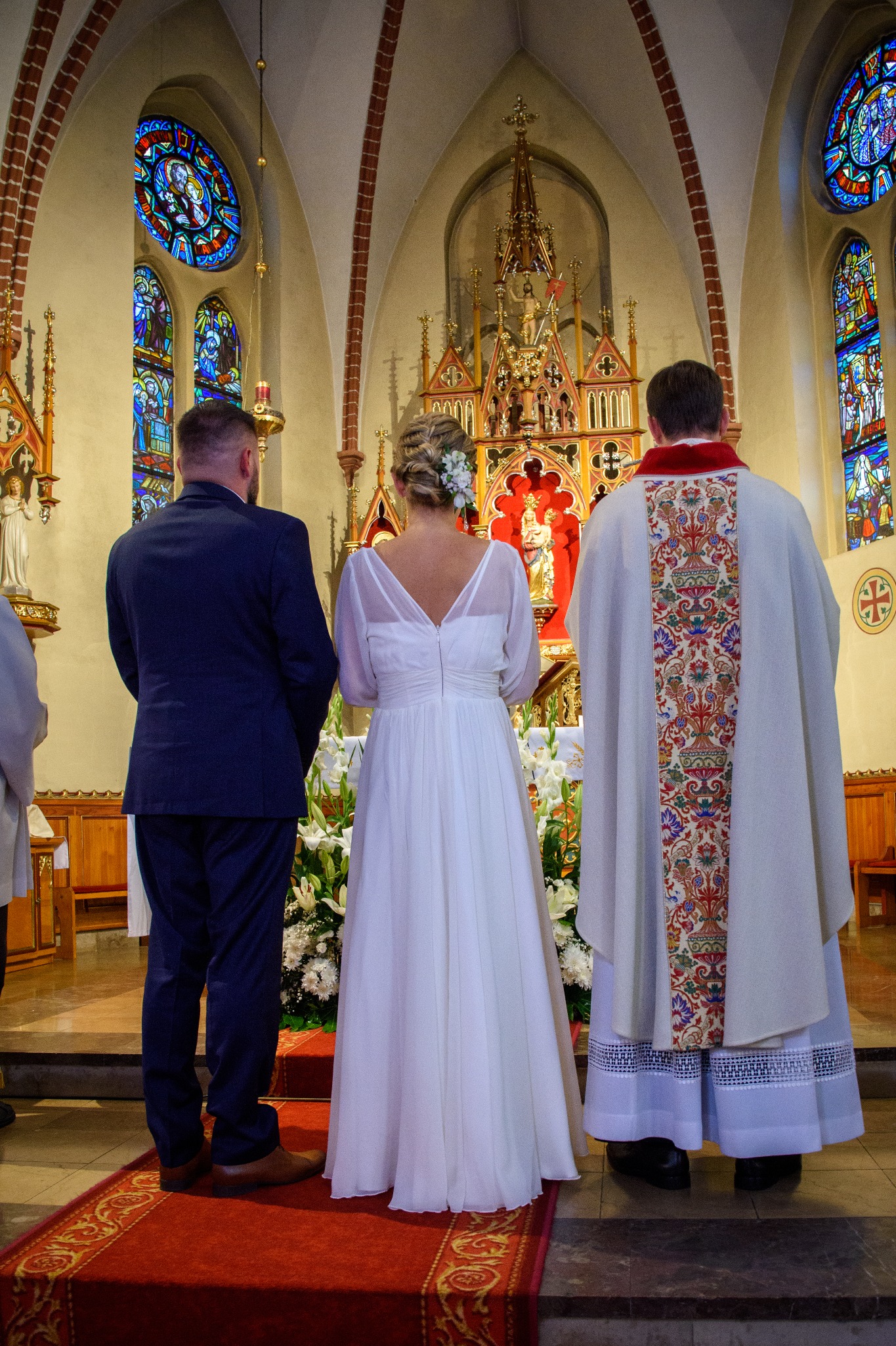 Para młoda i ksiądz stoją przed ołtarzem w kościele, widok z tyłu, na głowie panny młodej wpięte białe kwiaty, w tle witraże i bogato zdobiony ołtarz.