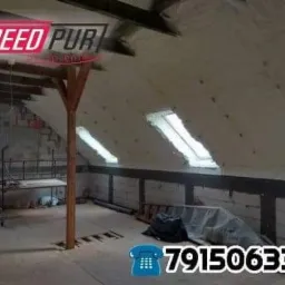 Poddasze w trakcie izolacji, widoczne okna dachowe, drewniana konstrukcja i świeżo nałożona warstwa ocieplenia. Na podłodze widoczne narzędzia i materiały budowlane.