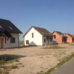 Nowe domy jednorodzinne w trakcie budowy: białe elewacje, ciemne dachy z oknami dachowymi, ceglane budynki w tle, częściowo wykończone, widoczny samochód dostawczy.