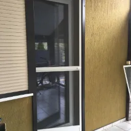 Otwarta moskitiera ramkowa w drzwiach balkonowych, z widoczną siatką i mechanizmem blokującym, zamontowana obok opuszczonej rolety zewnętrznej w kolorze beżowym.