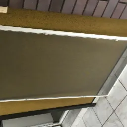 Biała rama okienna z siatką przeciw owadom, zamocowana na żółtej ścianie z widocznymi elementami mocującymi, ujęcie z dołu.