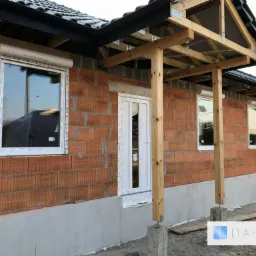 Surowy budynek w trakcie montażu stolarki okiennej i drzwiowej, widoczne okna z zamontowanymi roletami zwiniętymi w kasecie, białe drzwi wejściowe oraz drewniana konstrukcja zadaszenia nad wejściem.