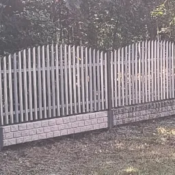 Metalowe ogrodzenie panelowe z podmurówką imitującą cegłę, oddzielające posesję od zieleni. Pionowe sztachety z zaokrąglonym wykończeniem.