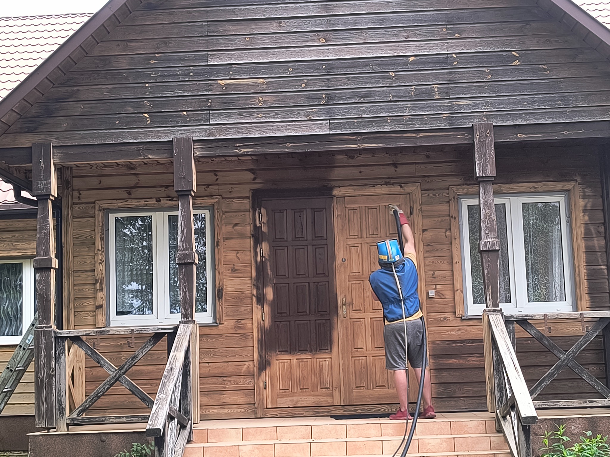 Pracownik w kombinezonie ochronnym maluje drewniany dom metodą natryskową, widoczna różnica w kolorze między pomalowaną a niepomalowaną częścią elewacji i drzwi.