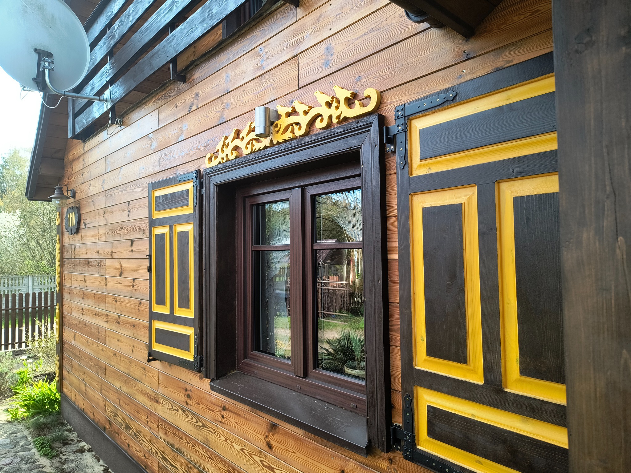 Drewniany dom z oknem i okiennicami w kolorach brązu i żółci, ozdobiony dekoracyjnym elementem nad oknem. Widoczna antena satelitarna i fragment ogrodu.