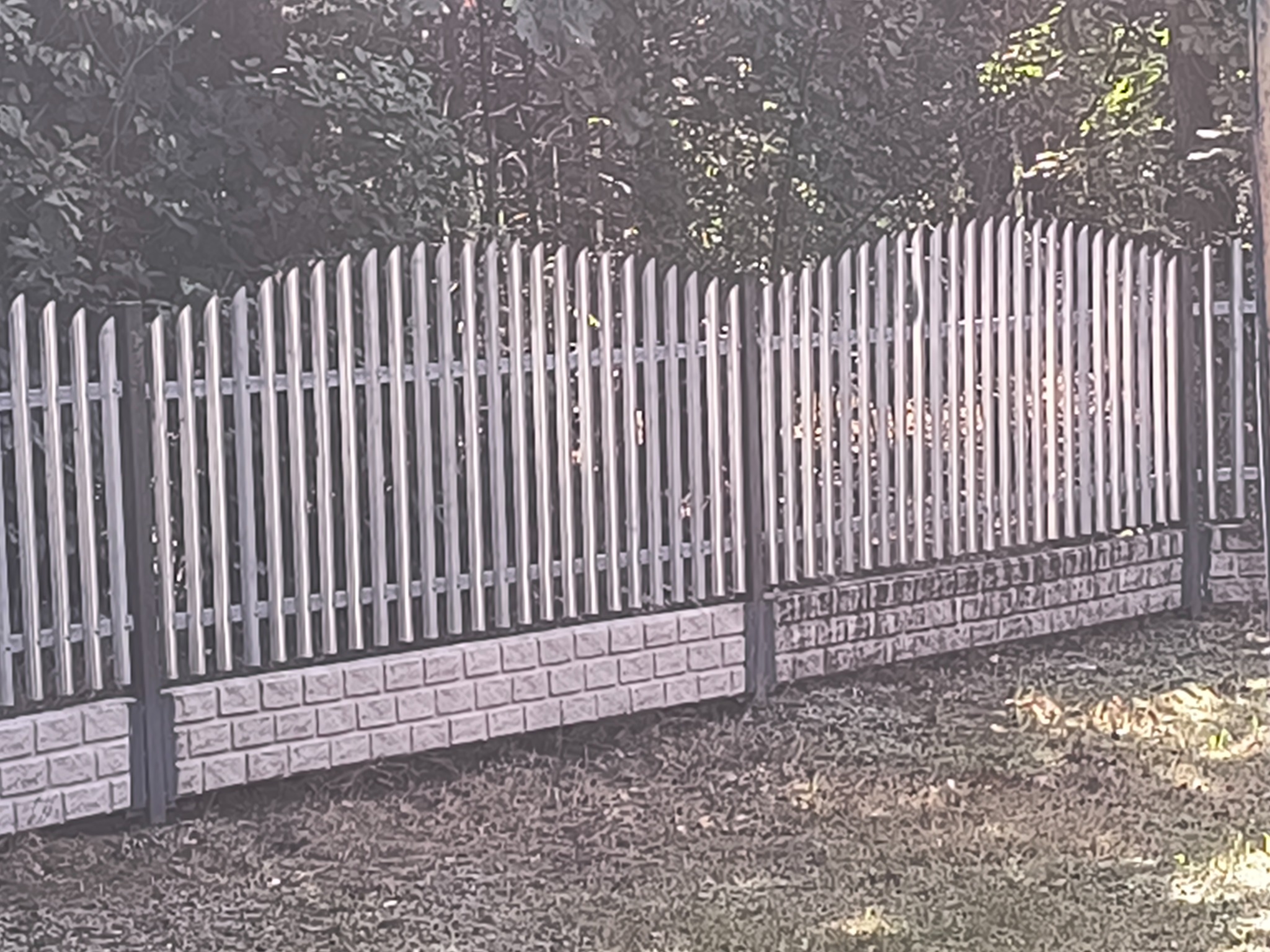 Metalowe ogrodzenie panelowe z podmurówką imitującą cegłę, oddzielające posesję od zieleni. Pionowe sztachety z zaokrąglonym wykończeniem.