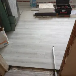 Jasne panele podłogowe ułożone w pokoju, widoczny próg z poziomnicą, narzędzia w skrzynce, paczki paneli i worek z zaprawą.