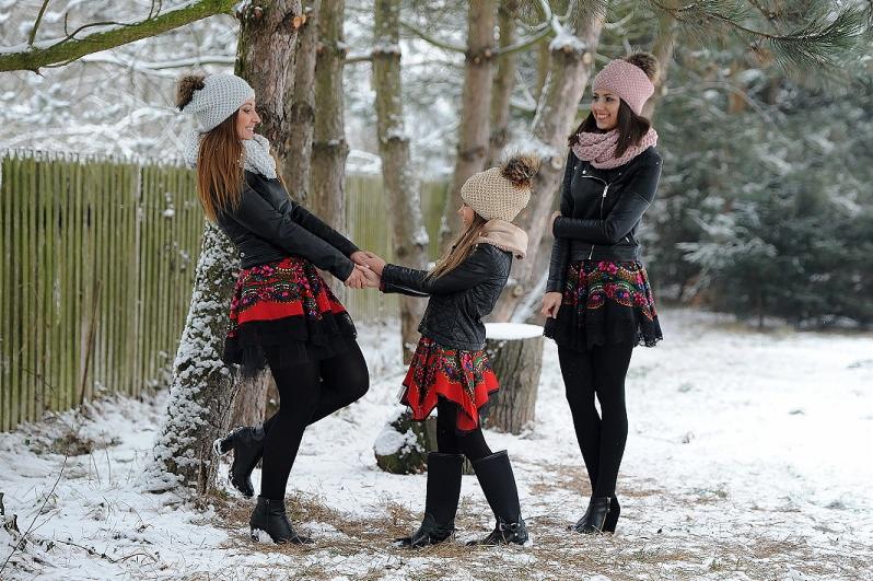 Trzy kobiety w zimowej scenerii, ubrane w czarne ramoneski, wzorzyste spódnice z folkowymi motywami i dopasowane czapki z pomponami, trzymające się za ręce na tle ośnieżonych drzew.