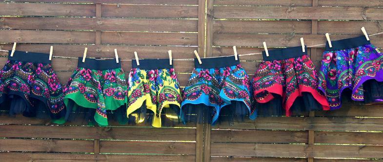 Siedem kolorowych, wzorzystych spódniczek z czarnym tiulem, suszących się na sznurku przypiętych klamerkami na tle drewnianego płotu.