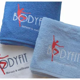 Stos trzech ręczników: ciemnoniebieski, jasnoniebieski i biały, każdy z wyhaftowanym logo 'BODYFIT fitness & wellness' z czerwoną sylwetką tancerza.