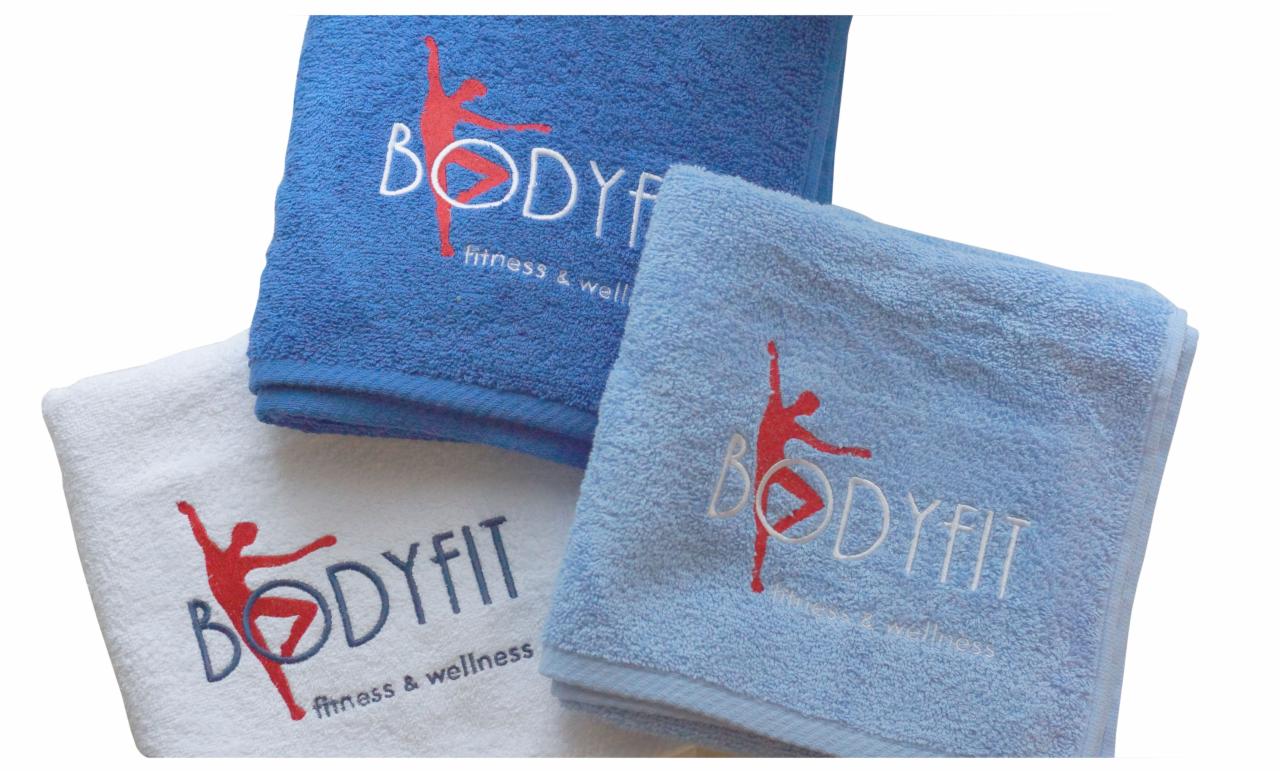 Stos trzech ręczników: ciemnoniebieski, jasnoniebieski i biały, każdy z wyhaftowanym logo 'BODYFIT fitness & wellness' z czerwoną sylwetką tancerza.