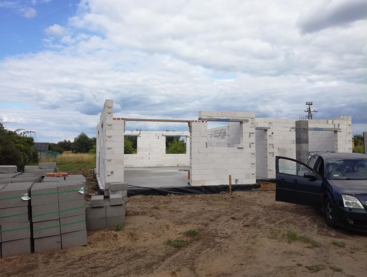 Wznoszone ściany budynku z białych bloczków, widoczny fundament, stos bloczków obok, oraz otwarte drzwi samochodu osobowego na pierwszym planie.