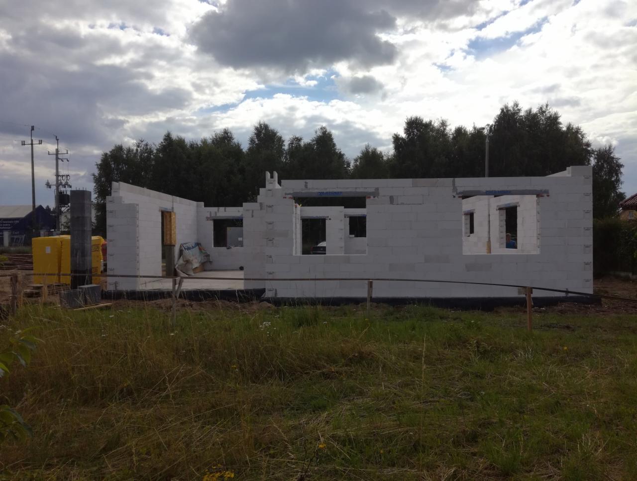 Wznoszenie ścian z białych bloczków na budowie domu jednorodzinnego, widoczne otwory okienne i drzwiowe, w tle drzewa i pochmurne niebo.