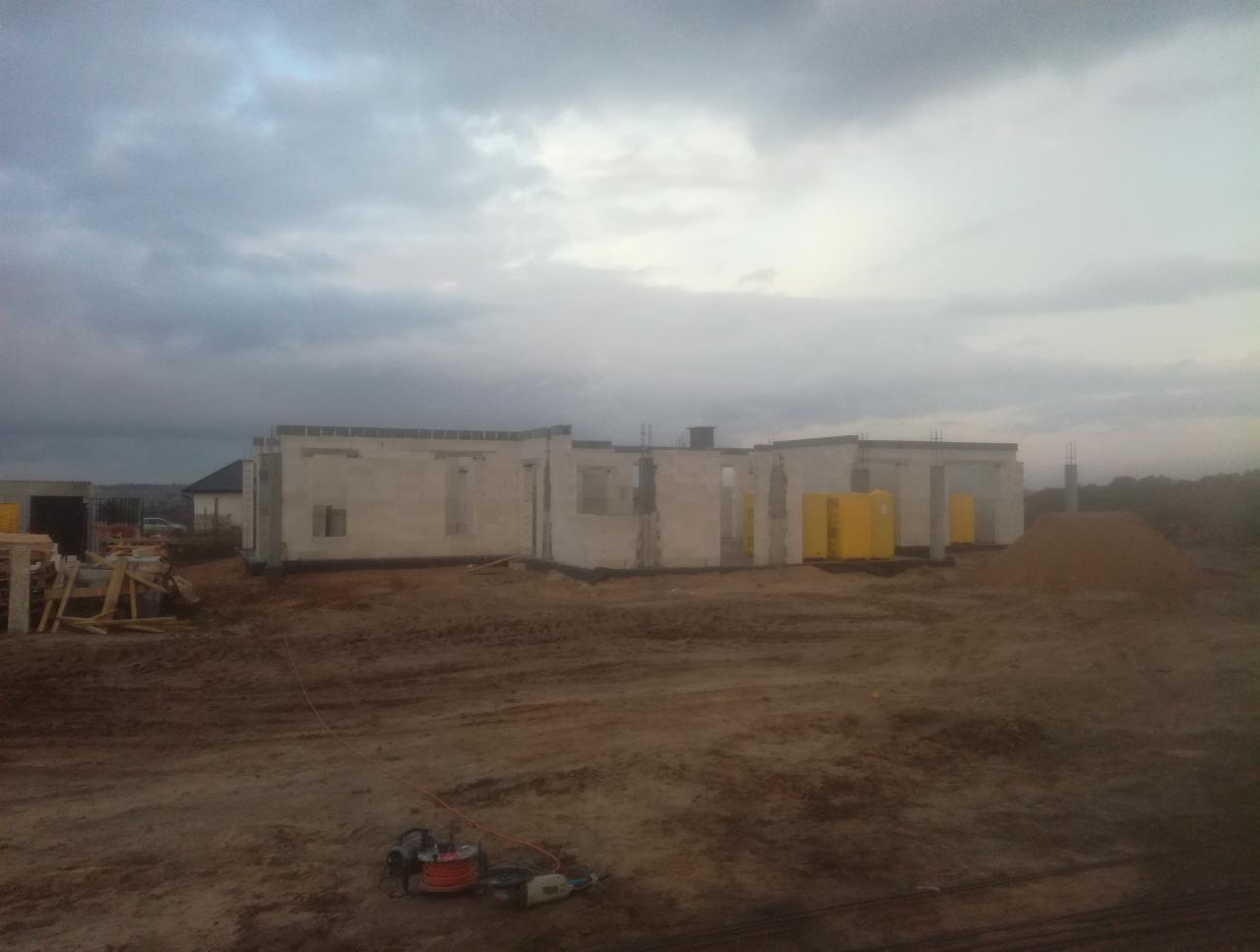 Wznoszone ściany domu z betonu komórkowego, widoczne żółte bloczki izolacyjne i narzędzia budowlane na ziemi, pochmurne niebo w tle.