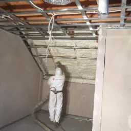 Pracownik w kombinezonie ochronnym aplikuje pianę otwartokomórkową na poddaszu, widoczna konstrukcja metalowa sufitu i instalacje.