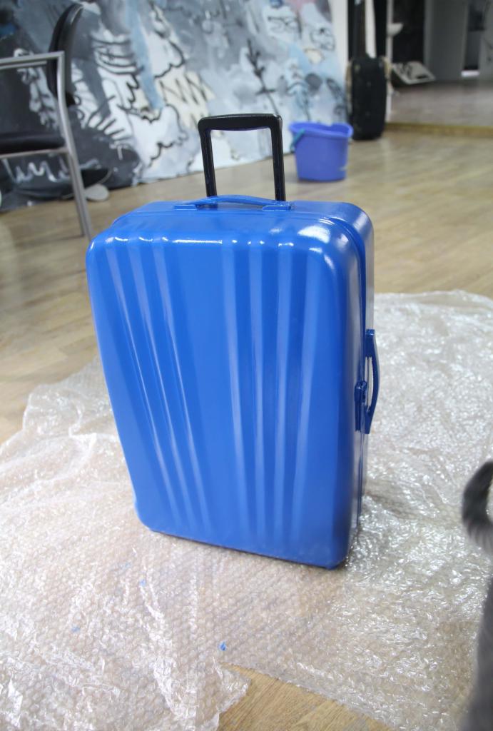 Błękitna walizka z wysuwaną rączką, zabezpieczona folią bąbelkową, ustawiona na podłodze z jasnego drewna, w tle widoczny malowany obraz abstrakcyjny i krzesło.