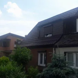 Szeregówka domów jednorodzinnych z brązowymi dachami, widoczne okna dachowe i rynny, fragmenty elewacji i zieleni.