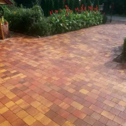 Kolorowa nawierzchnia z kostki brukowej w odcieniach czerwieni i żółci, z widoczną studnią z daszkiem i rabatą z czerwonymi kwiatami w tle.