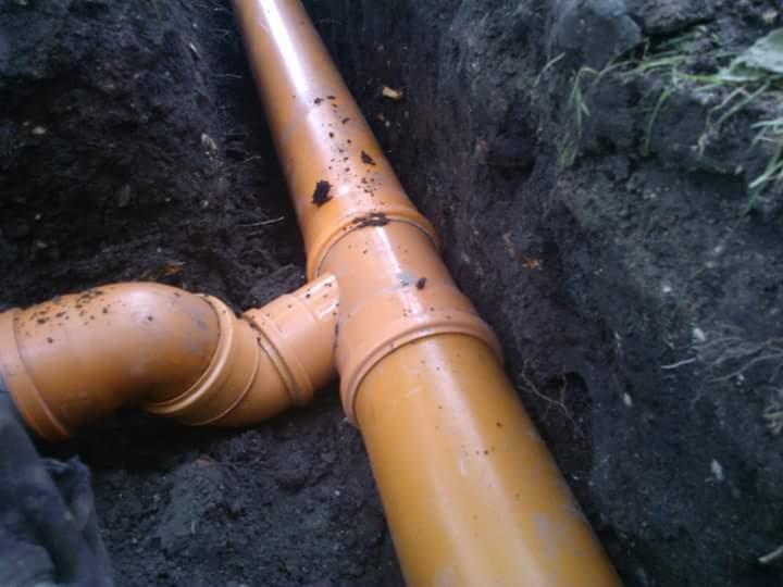 Pomarańczowe rury kanalizacyjne ułożone w wykopie, widoczne złącza i ciemna ziemia wokół.