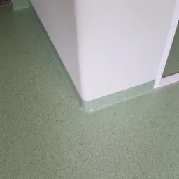 Fragment podłogi z zielonej, drobnoziarnistej wykładziny PCV przy białej ścianie z zaokrąglonym cokołem, widoczne łączenie wykładziny z cokołem.