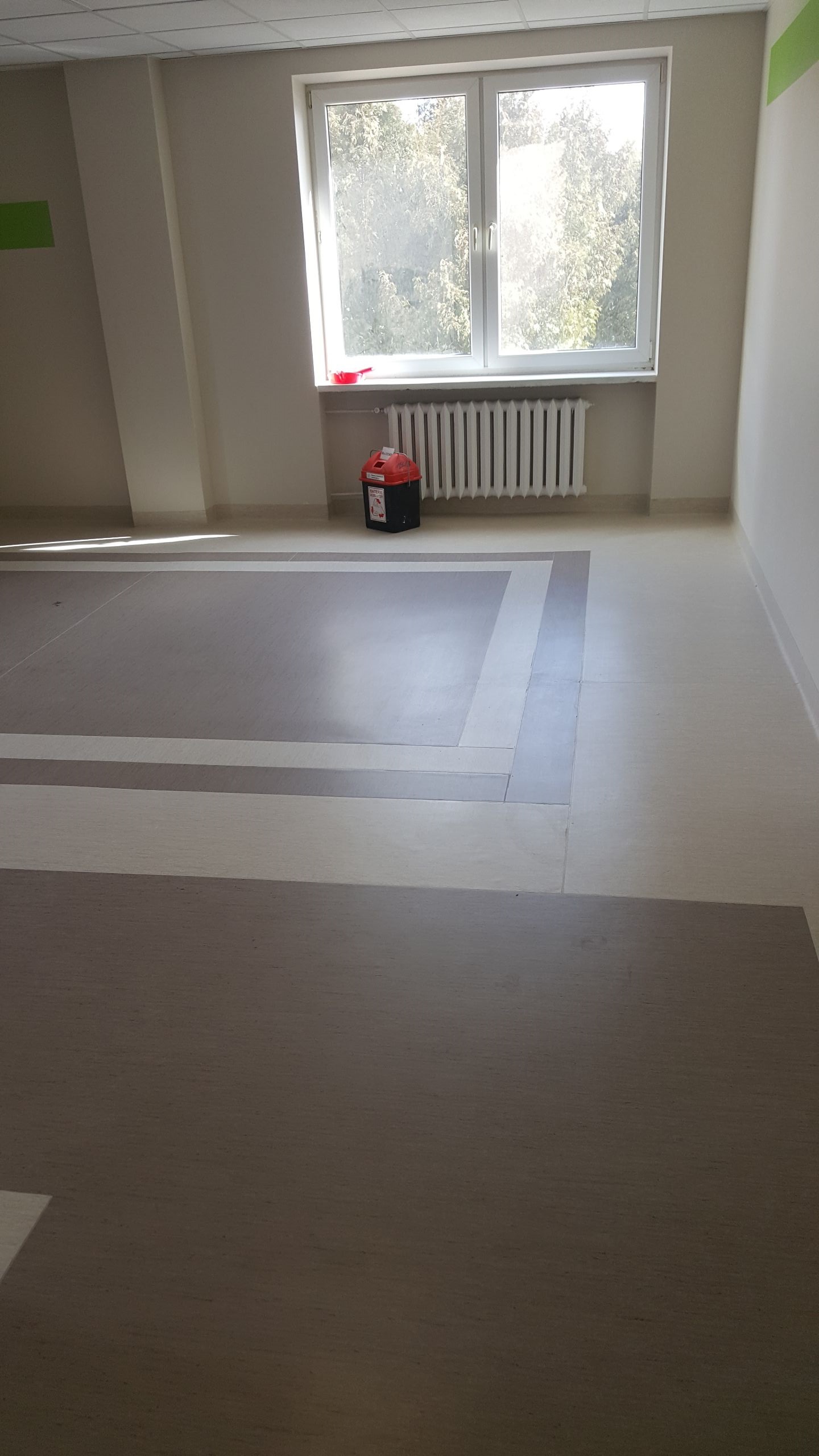 Wnętrze pomieszczenia z wykończoną podłogą w odcieniach beżu i szarości, widoczny grzejnik pod oknem i pojemnik na odpady.