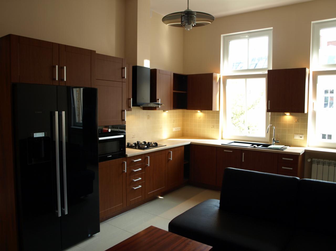 Nowoczesna kuchnia z brązowymi szafkami, czarną lodówką side-by-side z dystrybutorem wody, piekarnikiem w zabudowie, płytą gazową i czarnym okapem, oświetlona halogenami podszafkowymi, widok...