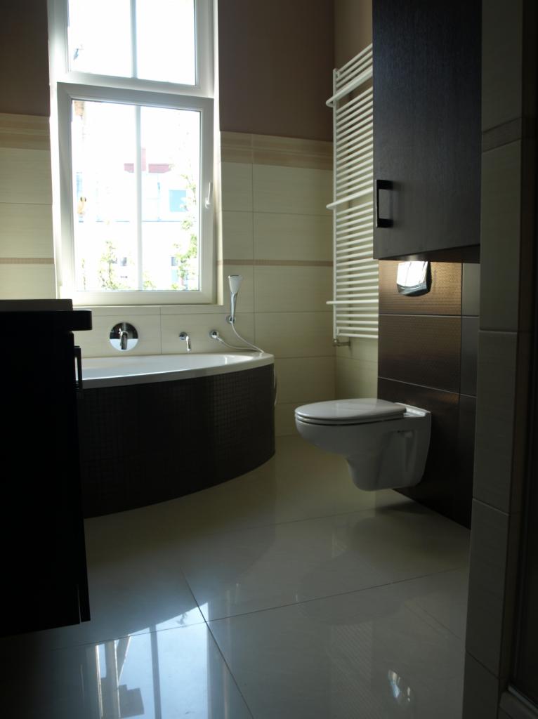 Nowoczesna łazienka z owalną wanną, podwieszaną toaletą i grzejnikiem drabinkowym, utrzymana w beżowo-brązowej kolorystyce z połyskującymi płytkami podłogowymi.