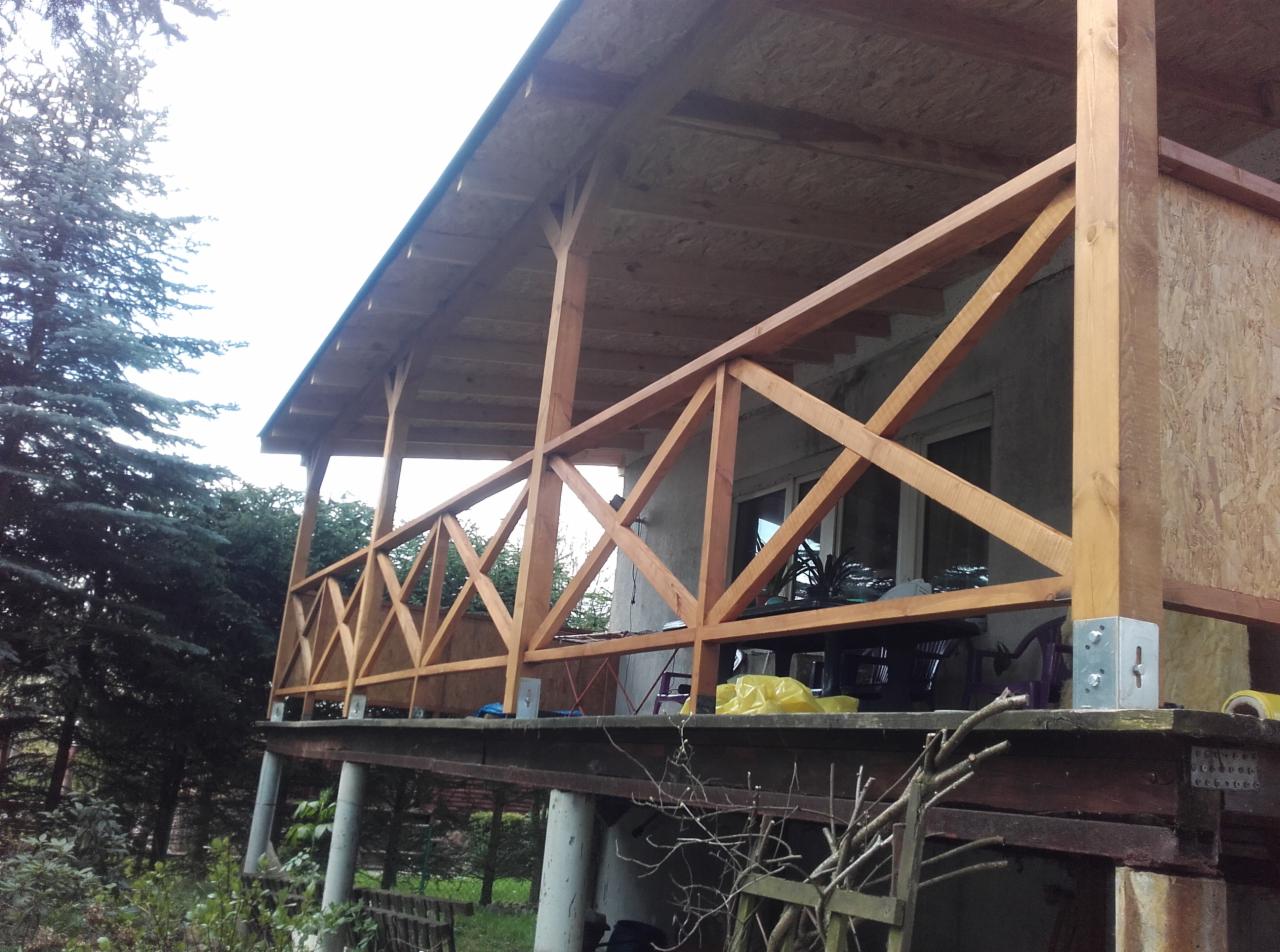 Zadaszony taras z drewnianą balustradą w geometryczny wzór, wsparty na betonowych słupach, widok z zewnątrz w pochmurny dzień.