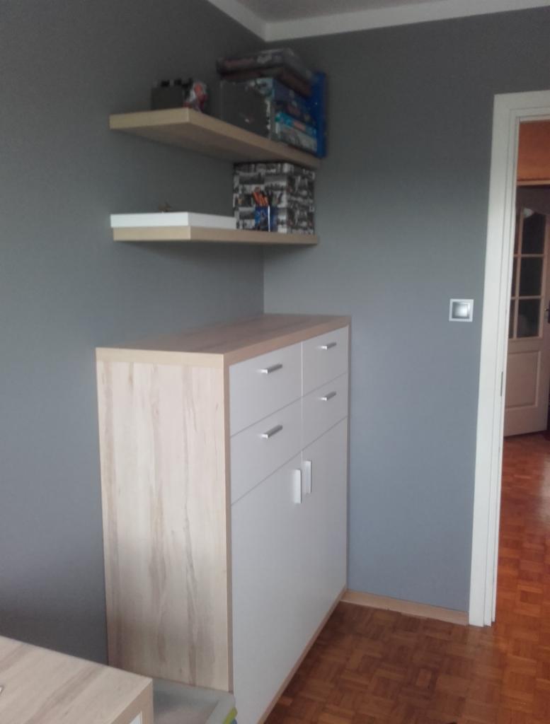 Szara ściana z dwiema półkami, na których stoją pudełka i gry, oraz biała komoda z czterema szufladami i dwojgiem drzwiczek. W tle widać otwarte drzwi i parkiet.