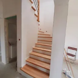 Drewniane schody o jasnych stopniach i czarnych metalowych balustradach w trakcie montażu, widoczne narzędzia i materiały budowlane na podłodze, surowe wykończenie ścian.