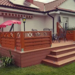 Przydomowy taras z kompozytu w kolorze brązowym, z balustradą z motywem geometrycznym, schodami z oświetleniem punktowym i meblami ogrodowymi na tle białego domu z czerwoną dachówką.