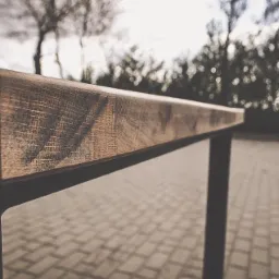 Fragment blatu drewnianego stołu z metalowymi nogami, widoczne słoje drewna i tekstura, na zewnątrz, w tle drzewa i kostka brukowa.