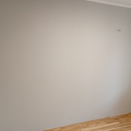 Firma Handlowo-Usługowa Marek Woźniak - Pomalowana na szaro ściana w rogu pokoju z parkietem na podłodze i wystającym kablem. Widoczne wykończenia sufitowe i fragment karnisza.