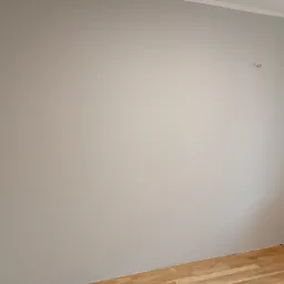 Pomalowana na szaro ściana w rogu pokoju z parkietem na podłodze i wystającym kablem. Widoczne wykończenia sufitowe i fragment karnisza.