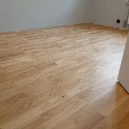 Jasne panele podłogowe ułożone w pokoju o szarych ścianach. Widoczny fragment białej futryny drzwi i grzejnik na ścianie. Ujęcie z niskiego kąta.