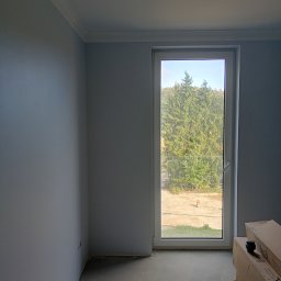 Firma Handlowo-Usługowa Marek Woźniak - Pomalowane na szaro wnętrze z oknem, widok na zewnątrz. Podłoga betonowa, przy oknie kartony. Listwa dekoracyjna pod sufitem. Nowe budownictwo.