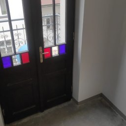 Firma Handlowo-Usługowa Marek Woźniak - Stylowe, ciemne drzwi wewnętrzne z kolorowymi szybkami w odcieniach czerwieni i granatu, widoczne na klatce schodowej. Charakterystyczne zdobienia szyb i widok na zewnątrz.