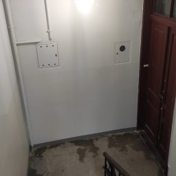 Firma Handlowo-Usługowa Marek Woźniak - Klatka schodowa po odświeżeniu: białe ściany z instalacjami, brązowe drzwi, schody z metalową barierką. Widoczne ślady wilgoci na podłodze.