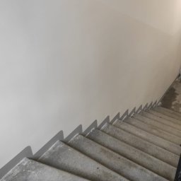 Firma Handlowo-Usługowa Marek Woźniak - Świeżo pomalowana ściana klatki schodowej w jasnym, neutralnym odcieniu szarości, widoczna z góry. Betonowe schody z czarną balustradą.