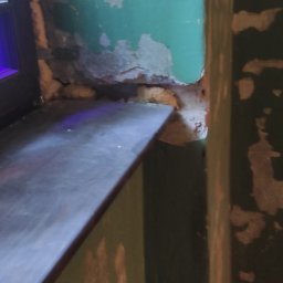 Firma Handlowo-Usługowa Marek Woźniak - Narożnik pokoju z oknem, ściany z odpryskującą farbą w odcieniach zieleni i beżu, ukazujące surowy stan przed remontem. Widoczne uszkodzenia i brak wykończenia.