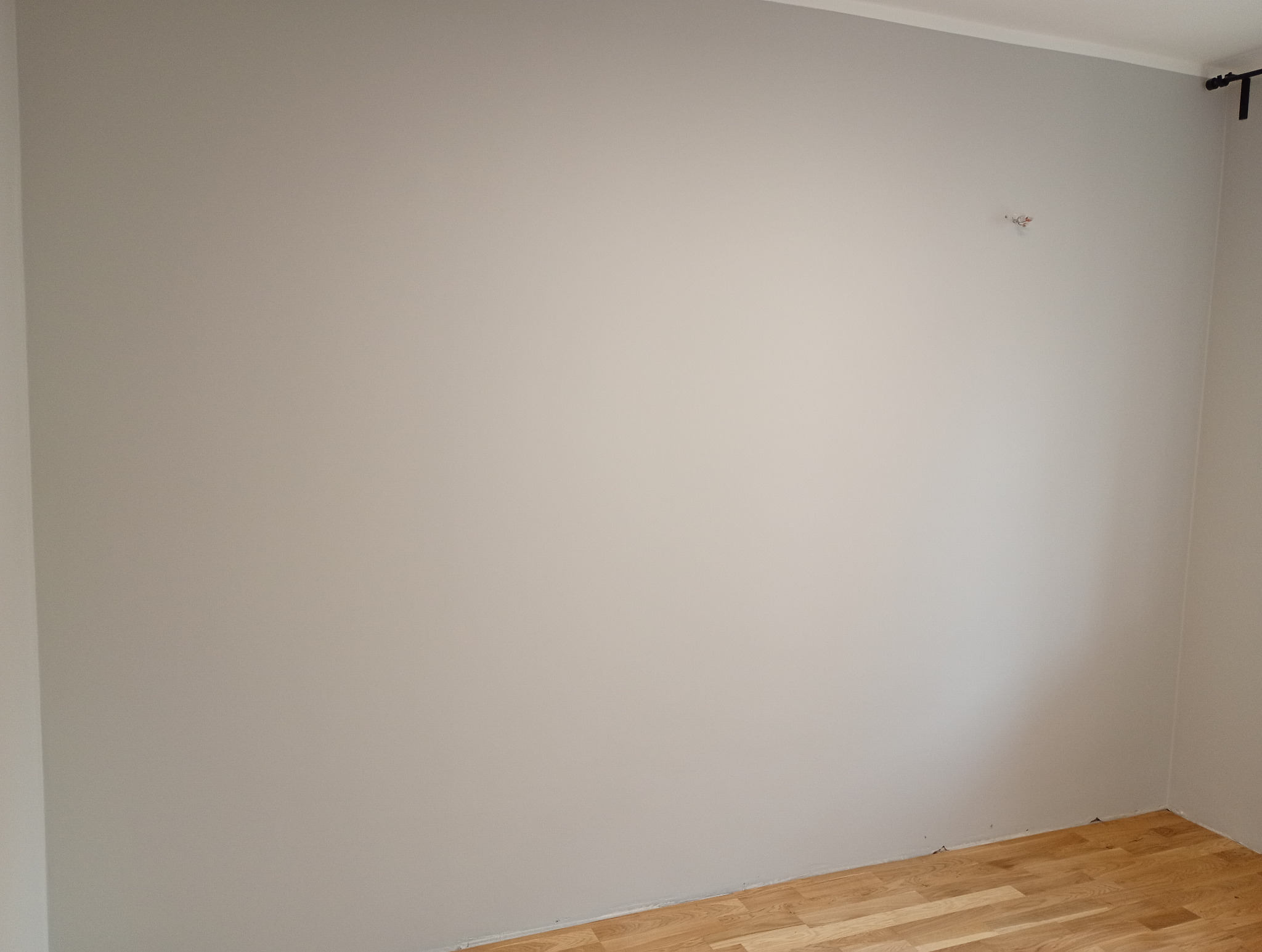 Pomalowana na szaro ściana w rogu pokoju z parkietem na podłodze i wystającym kablem. Widoczne wykończenia sufitowe i fragment karnisza.
