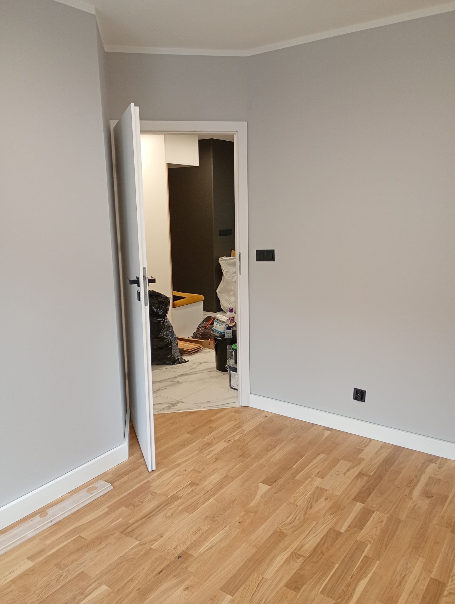 Szare ściany, drewniana podłoga i otwarte drzwi ukazujące wnętrze z pojemnikami i workiem. Widoczne gniazdka elektryczne. Wykończenie wnętrza w neutralnej kolorystyce.