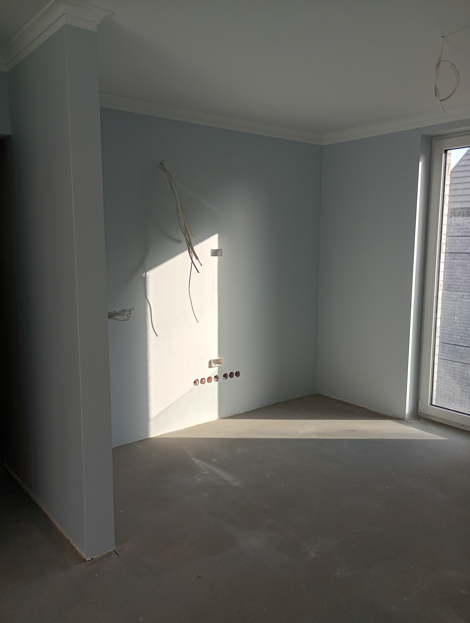 Jasne wnętrze po malowaniu ścian na pastelowy odcień, z widocznymi wyprowadzonymi kablami elektrycznymi i listwami przypodłogowymi, wpadające światło słoneczne.