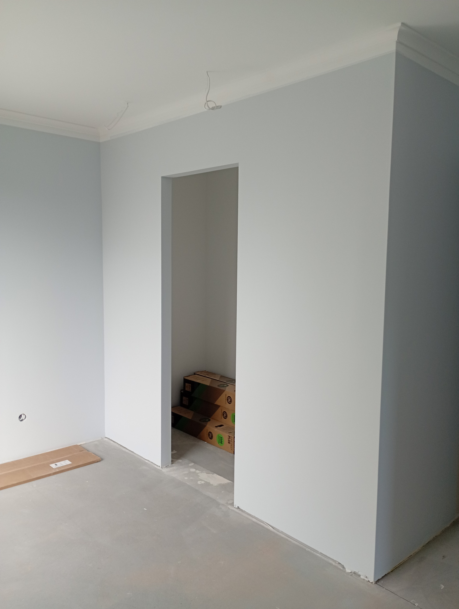 Jasne, pomalowane na szaro ściany z białą listwą przysufitową w nowym wnętrzu. Widoczne kable od oświetlenia, surowa podłoga i kartony w przejściu.
