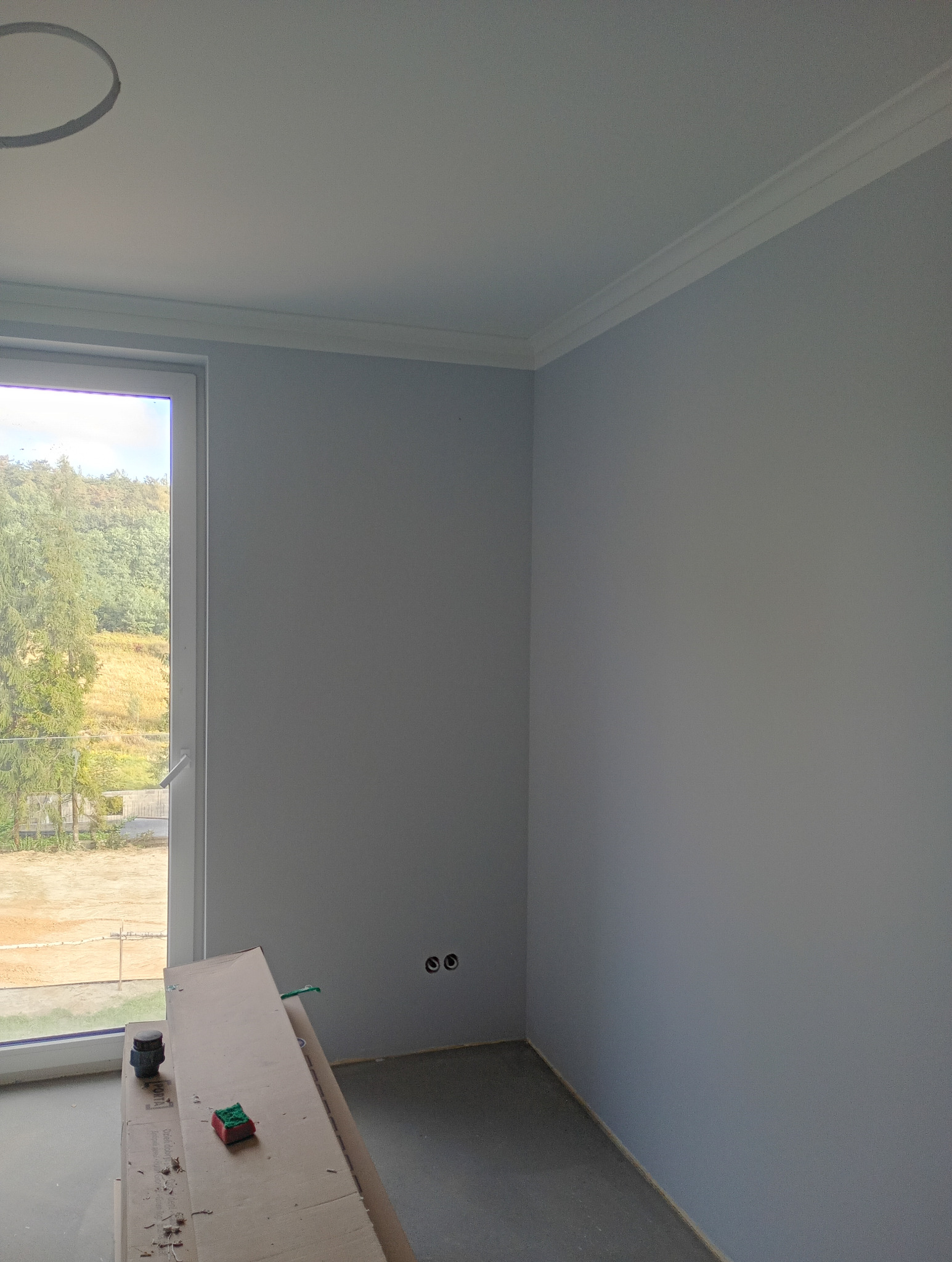Pomieszczenie po malowaniu na szaro, widoczne listwy przypodłogowe, okno z widokiem na zieleń, kartony na podłodze i gniazdka elektryczne.