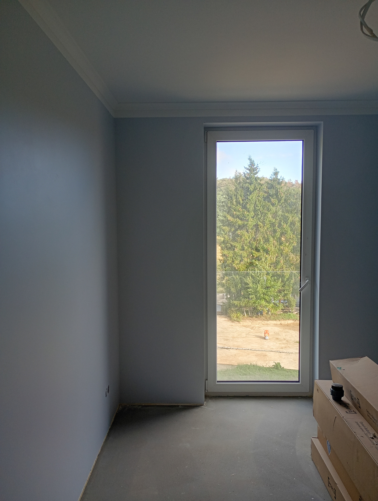 Pomalowane na szaro wnętrze z oknem, widok na zewnątrz. Podłoga betonowa, przy oknie kartony. Listwa dekoracyjna pod sufitem. Nowe budownictwo.