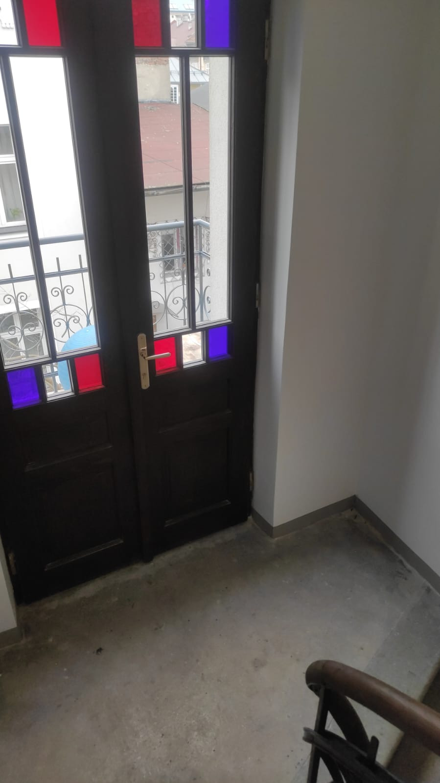 Stylowe, ciemne drzwi wewnętrzne z kolorowymi szybkami w odcieniach czerwieni i granatu, widoczne na klatce schodowej. Charakterystyczne zdobienia szyb i widok na zewnątrz.