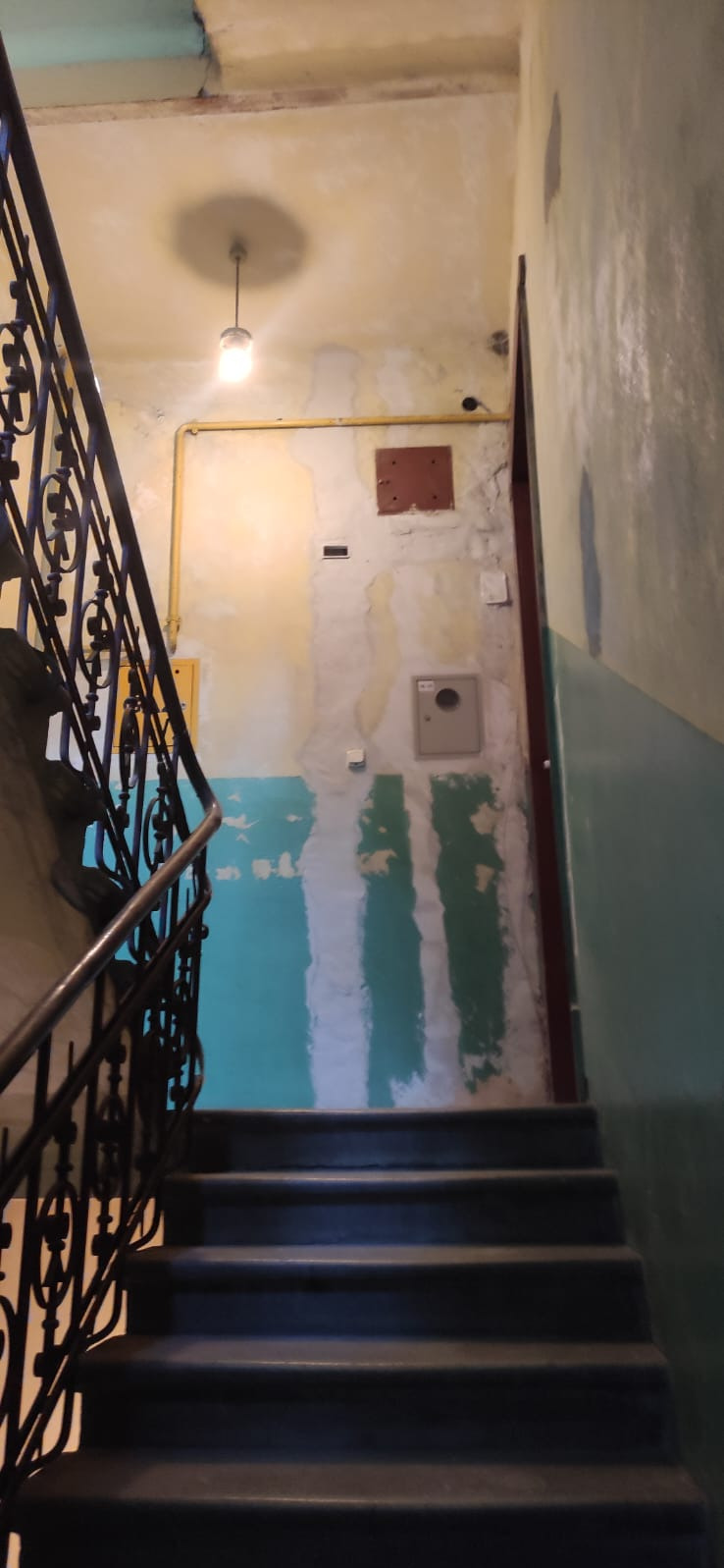 Klatka schodowa w trakcie remontu: widoczne fragmenty starej farby, przygotowane ściany do malowania, schody i metalowa balustrada. Oświetlenie sufitowe.