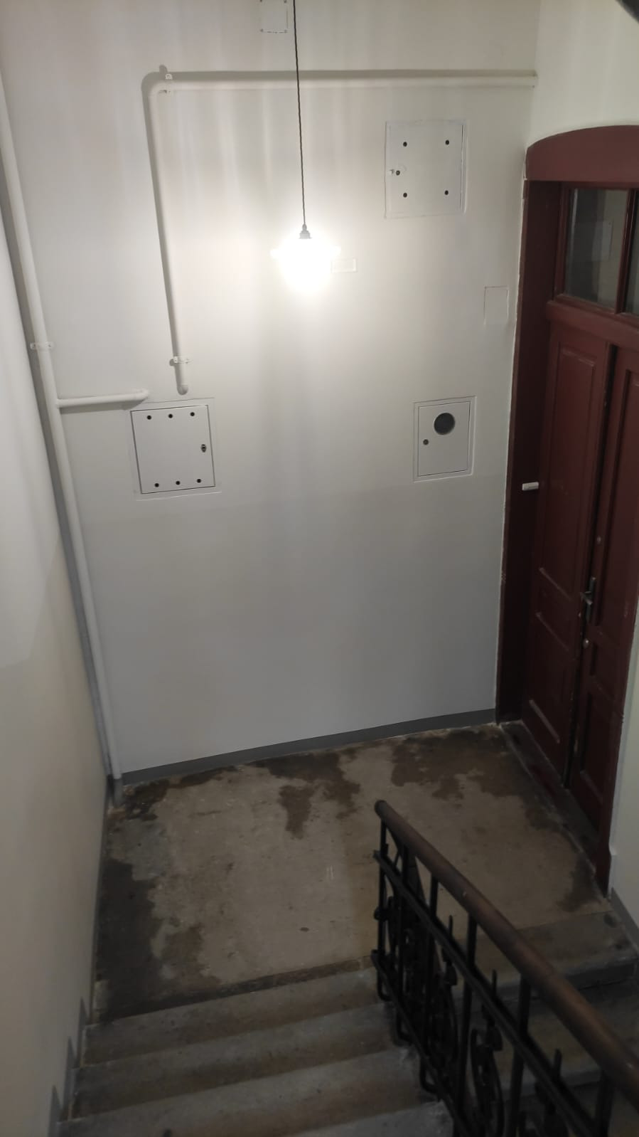 Klatka schodowa po odświeżeniu: białe ściany z instalacjami, brązowe drzwi, schody z metalową barierką. Widoczne ślady wilgoci na podłodze.